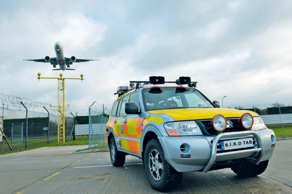 Car with speakers attached to roof at an airport with a plane flying over it